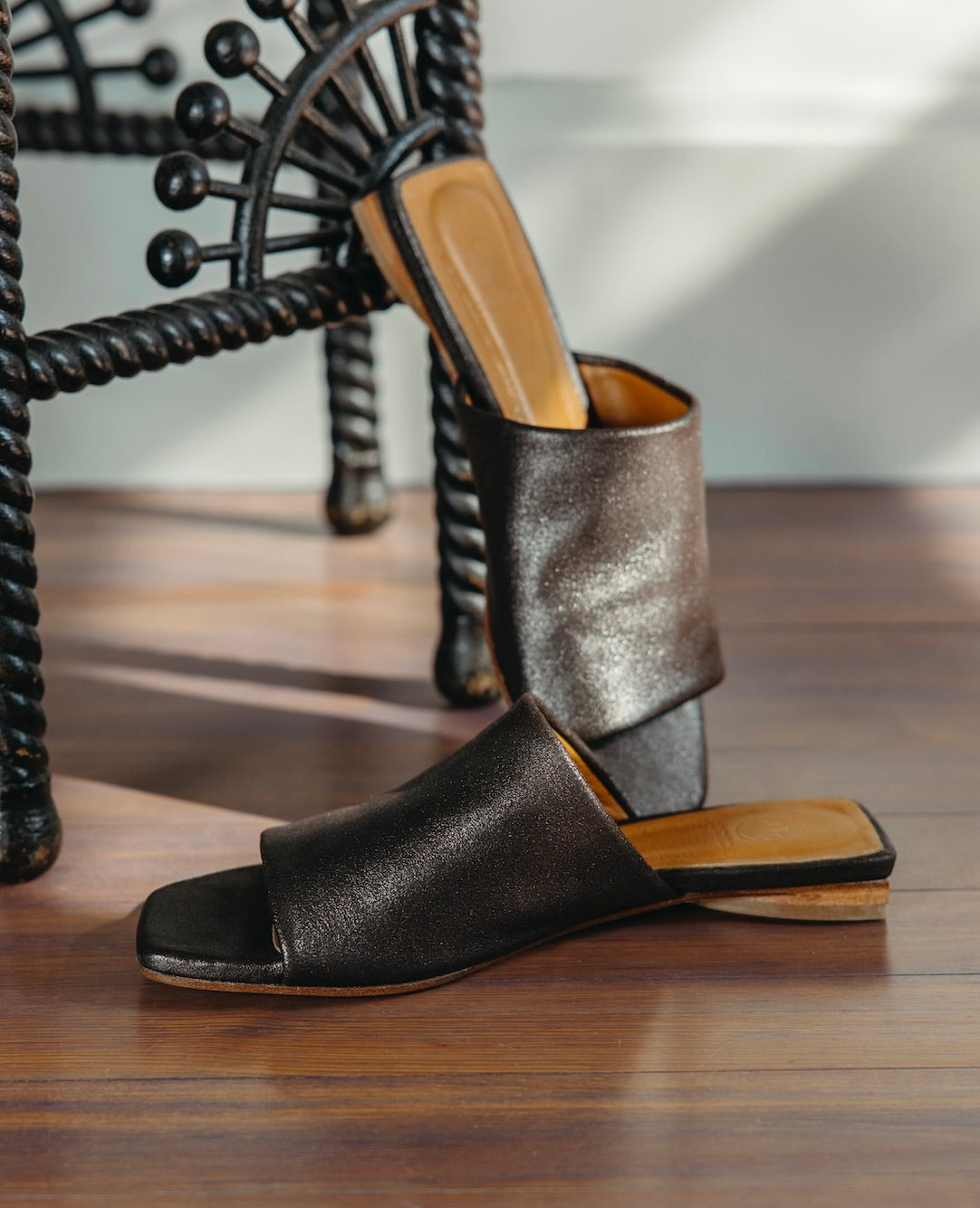 Close up view of the Ferhana Sandal in anthracite shimmer sued with the heel of the left sandal propped up against the night table.