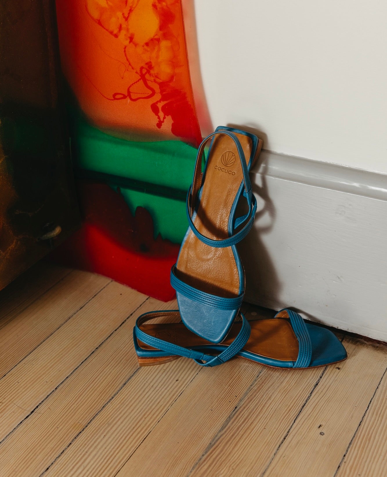The Fifi Sandal in Cerulean leather with the right pair propped up against the wall placed on top of the left pair. 