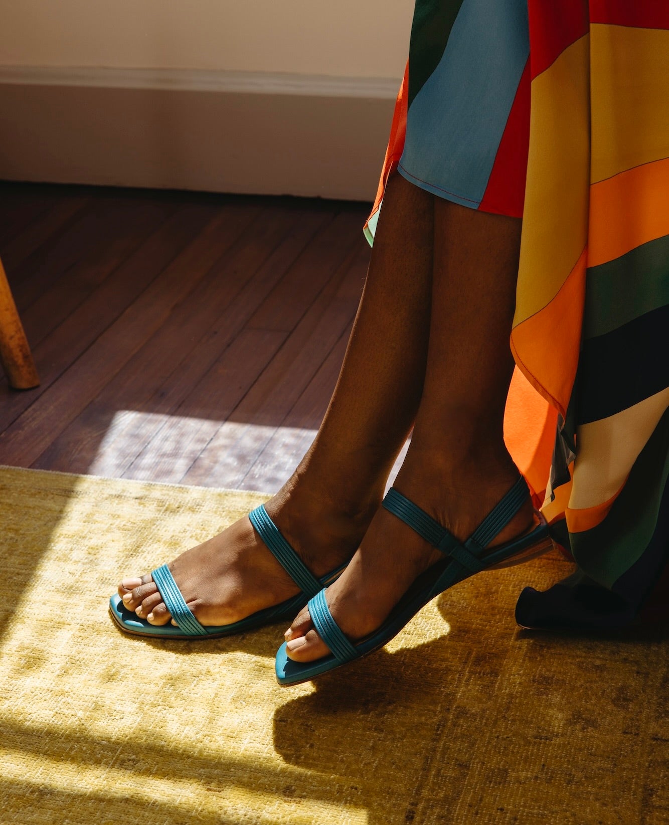 Feet crossed against a yellow rug wearing the Fifi Sandal in Cerulean leather. 