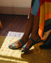 Feet crossed against a yellow rug wearing the Fifi Sandal in Cerulean leather.  6