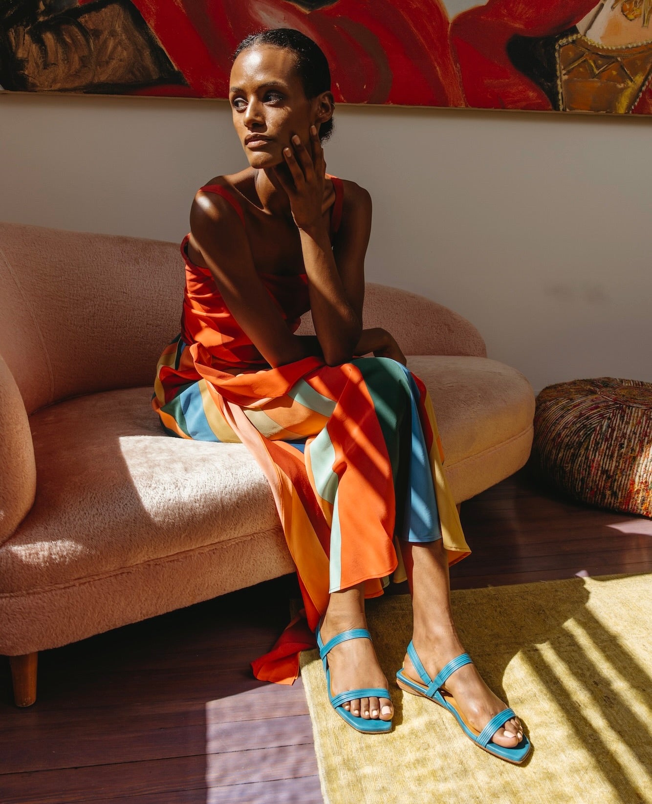Woman seated on a couch with legs crossed, glancing to the side, wearing  dress and the Fifi Sandal in Cerulean leather. 
