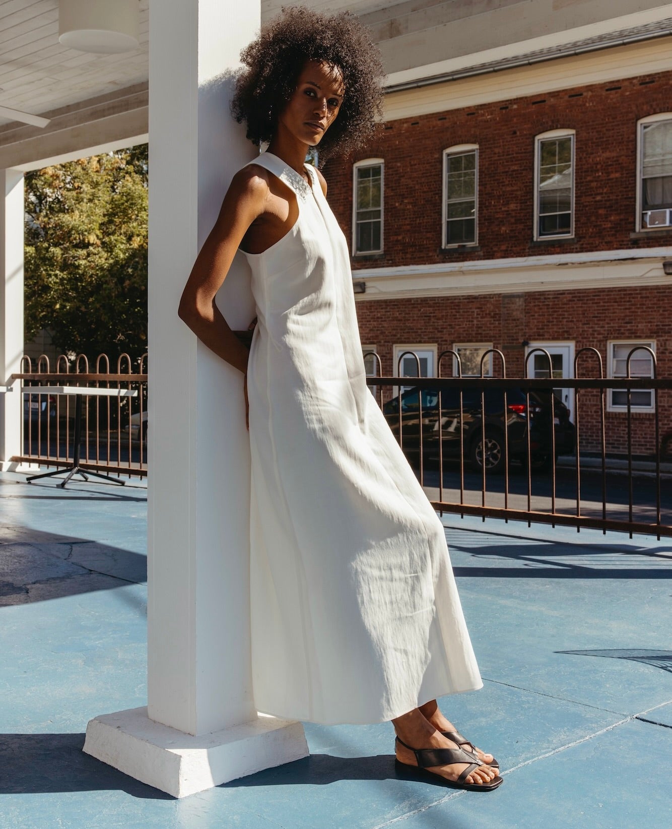 Woman wearing a white dress leaning against a white column wearing the Finch Sandal in Black. 