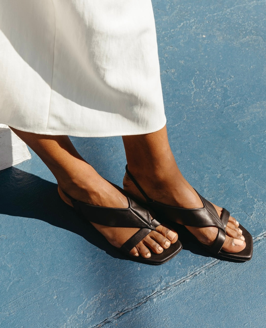 Feet placed on blue concrete flooring wearing the Finch Sandal in Black. 