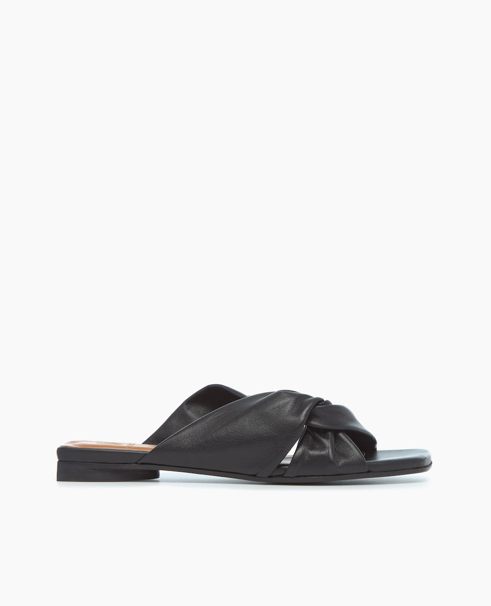 A black flat sandal featuring a sleek, minimalist design with a twisted knot detail on the strap, made from smooth leather material.