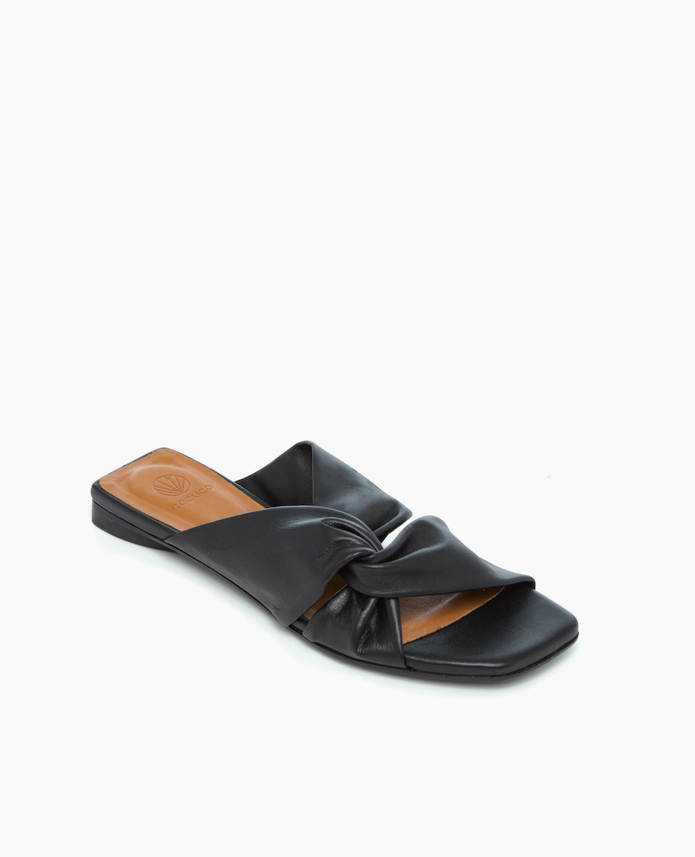 A sleek black leather slide sandal features a twisted knot design on the upper, a flat sole, and an open toe, with a smooth texture and a minimalist aesthetic.