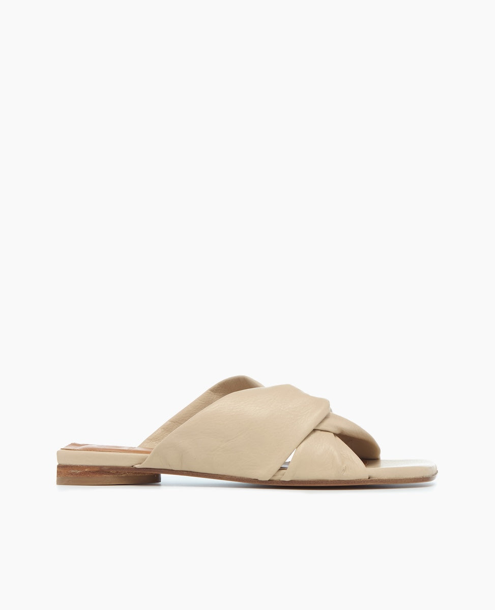 A beige, flat sandal with a minimalist design features a crisscross strap made of smooth leather and a thin, light brown sole.