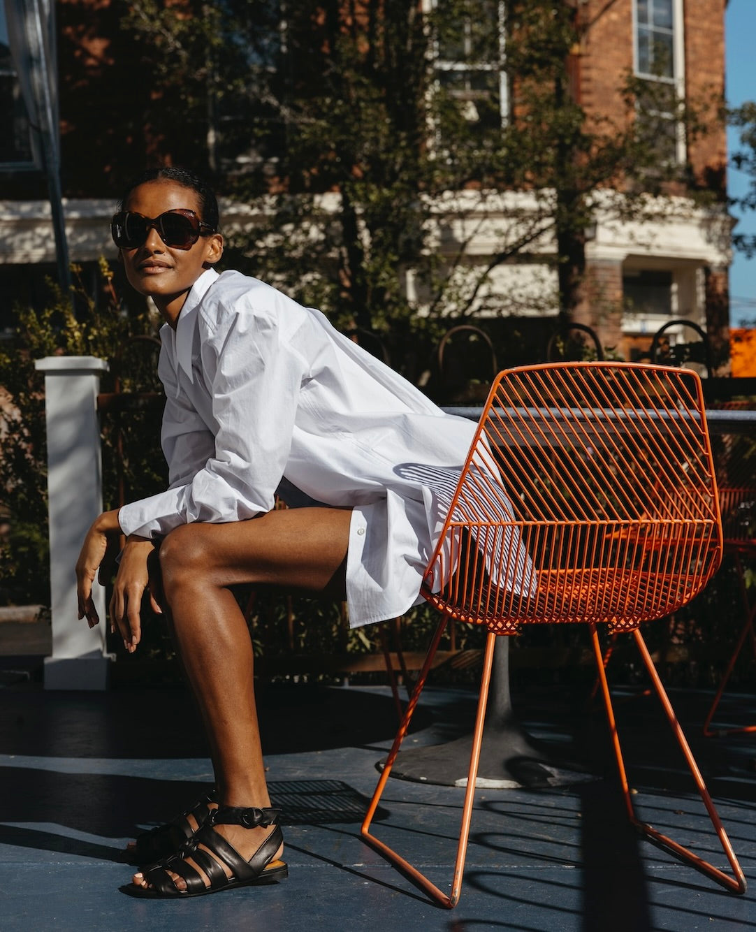 Woman wearing a white collared shirt and the Flair Sandal Black, sitting on orange chair leaning over. 