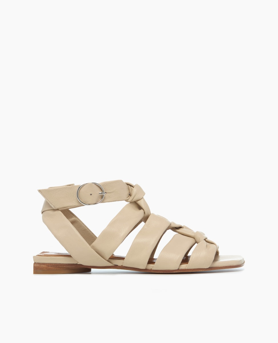 A beige sandal with a flat sole features multiple soft, padded straps crossing over the foot, a silver buckle on the ankle strap, and a smooth leather finish.