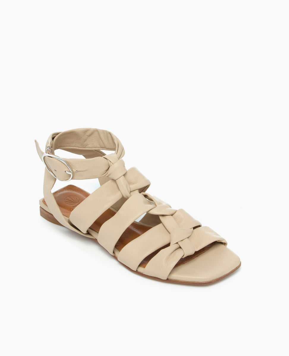 A beige sandal featuring a flat sole, multiple wide straps with knotted details, an ankle strap with a silver buckle, and a smooth leather texture.