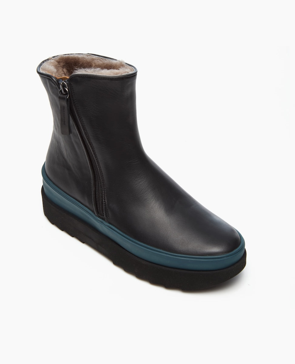 A sleek black leather ankle boot with a side zipper, featuring a plush fur lining, a thick teal midsole, and a textured black rubber outsole.