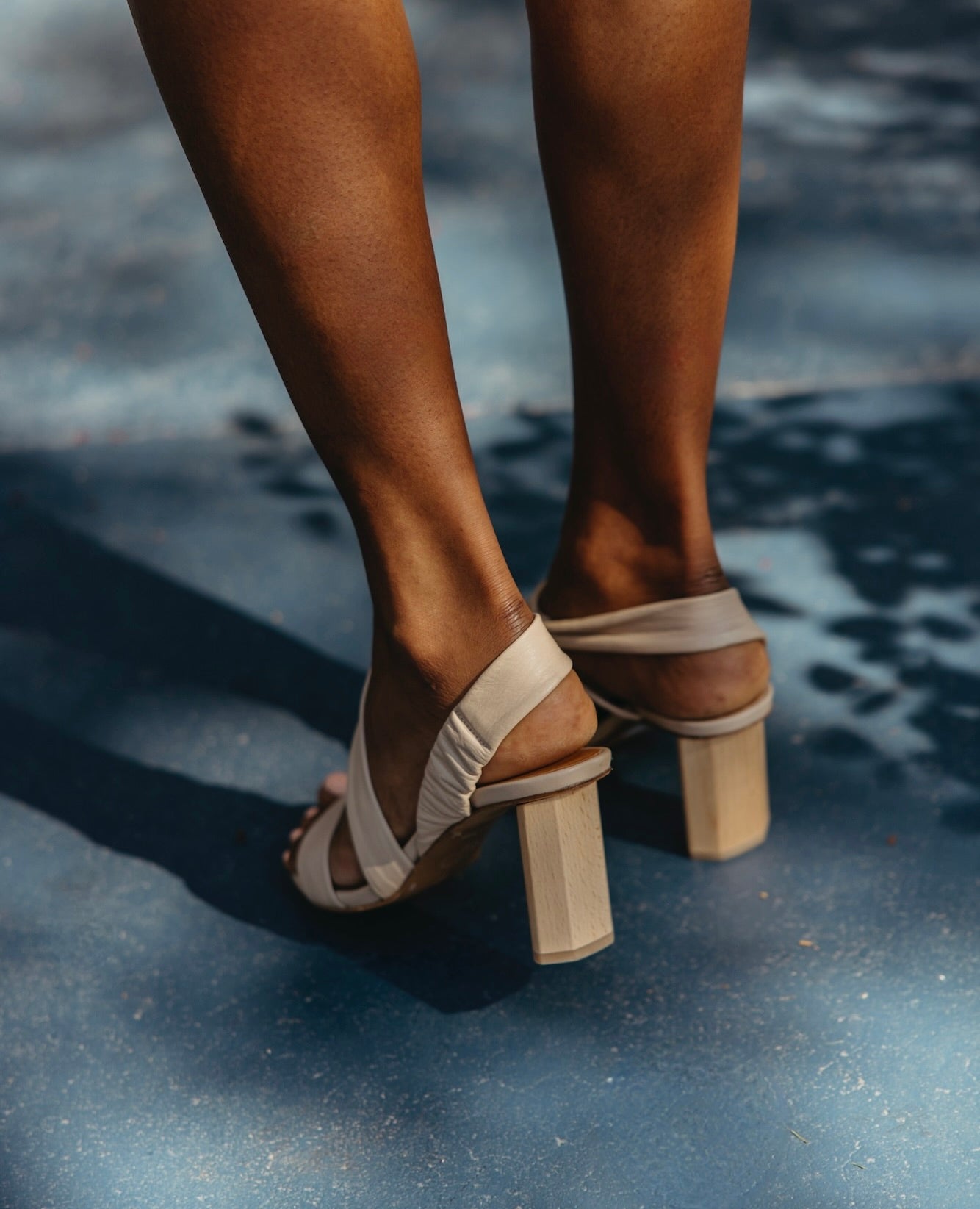 Behind the feet picture of legs and feet wearing the Gabby Heel in Latte Machiatto against a blue concrete floor. 