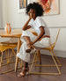 Woman seated on a yellow chair at a yellow table wearing white pants and top, the Gigi Heel in Fawn Nubuck. 2