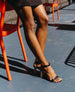 Legs of a woman wearing the Gina Heel in Black Pearl Patent leaning against an orange chair outside.  5