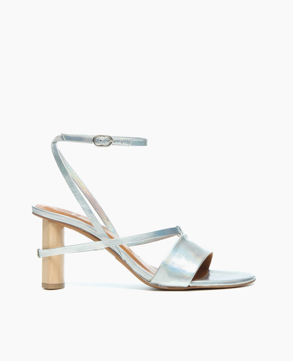 A sleek, metallic silver sandal with a thin ankle strap, a low wooden block heel, and a minimalist design featuring a single wide strap across the toes.