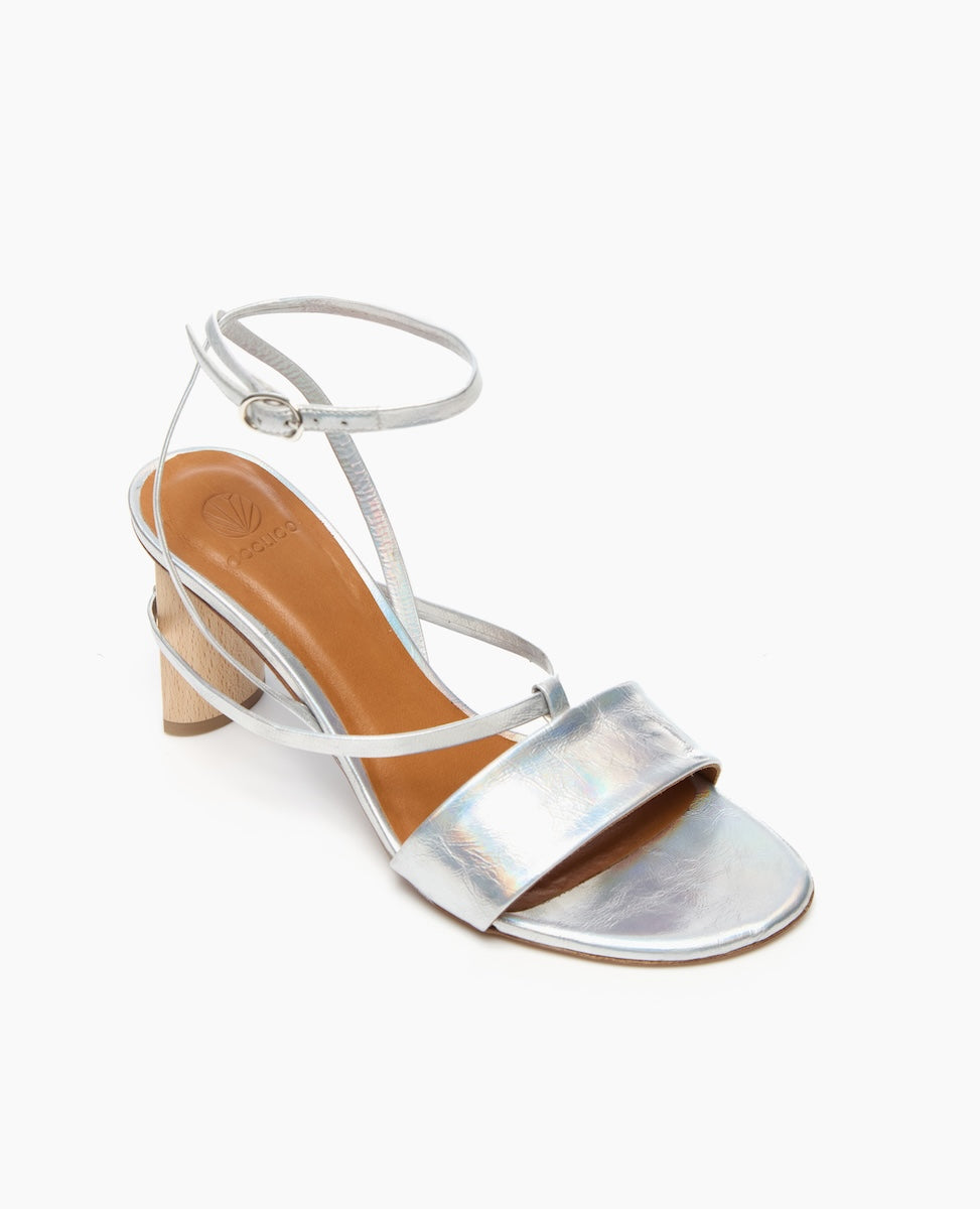A silver, open-toe sandal with a low wooden block heel, featuring a wide metallic strap across the front and a thin ankle strap with a buckle.