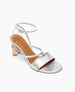 A silver, open-toe sandal with a low wooden block heel, featuring a wide metallic strap across the front and a thin ankle strap with a buckle. 6