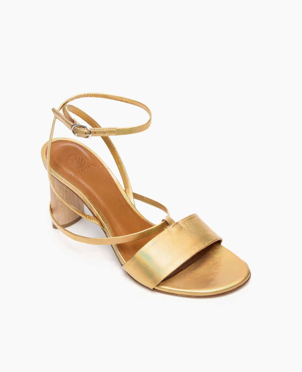 A gold sandal with a wooden wedge heel features a wide metallic strap across the toes, a thin ankle strap with a buckle, and a smooth, shiny texture.