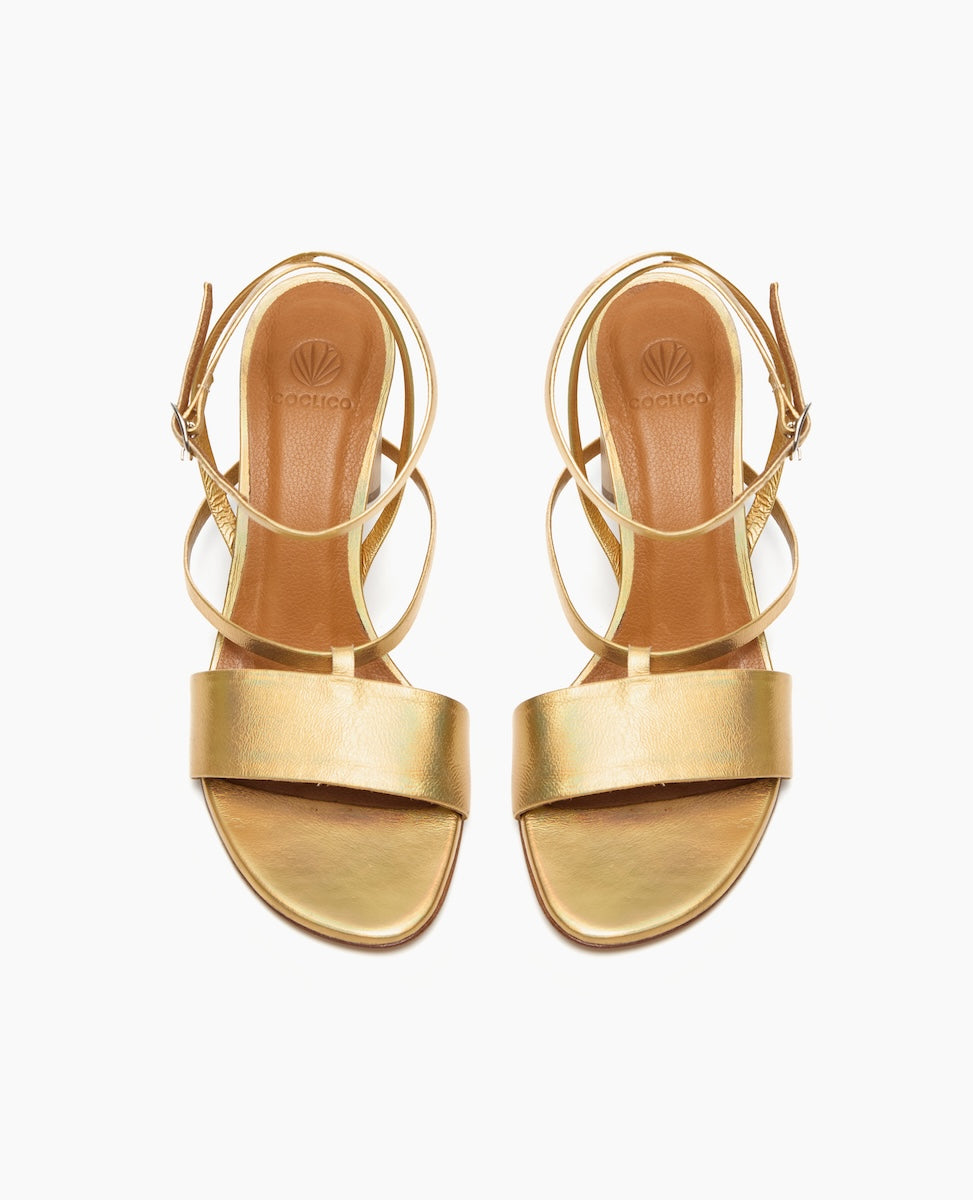 A pair of elegant, gold-colored sandals with a smooth, metallic finish, featuring a wide strap across the toes, a slender ankle strap with a buckle, and a flat sole with a subtle logo imprint on the insole.