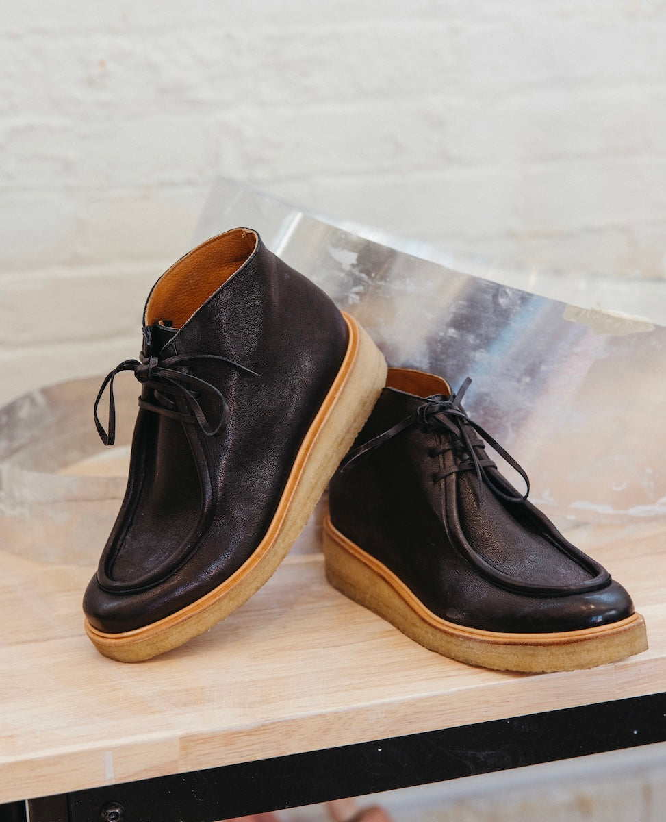 A pair of black leather ankle boots with a smooth texture, featuring lace-up fronts, softly sculpted toes, and light brown crepe rubber soles.