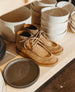 A pair of tan split suede boots with tan suede laces, featuring a softly sculpted toe and a light brown rubber sole, placed on a wooden shelf next to stacks of white and dark gray ceramic bowls and plates. 9