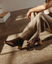 A person wearing ribbed, beige knit pants and metallic broze-colored ankle boots with a wooden block heel sits on a polished concrete floor. 11