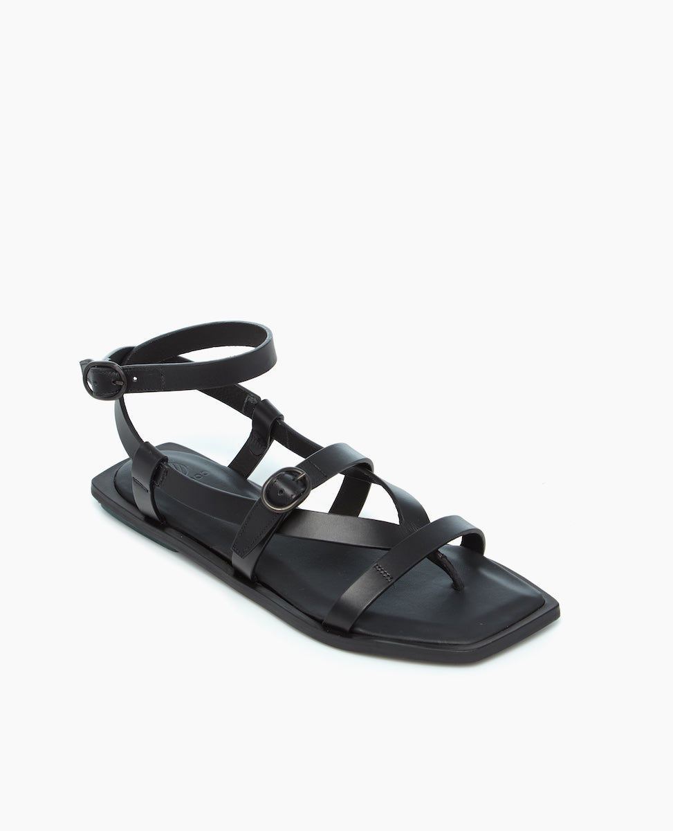 A black sandal featuring a flat sole, multiple thin straps crossing over the foot, an adjustable ankle strap with a buckle, and a sleek, minimalist design.