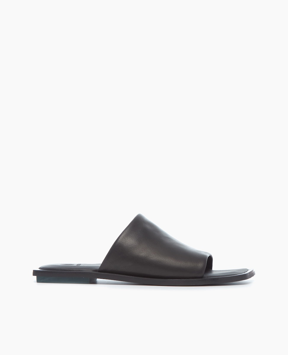A sleek, black leather slide sandal with a minimalist design, featuring a wide strap across the foot and a flat sole.