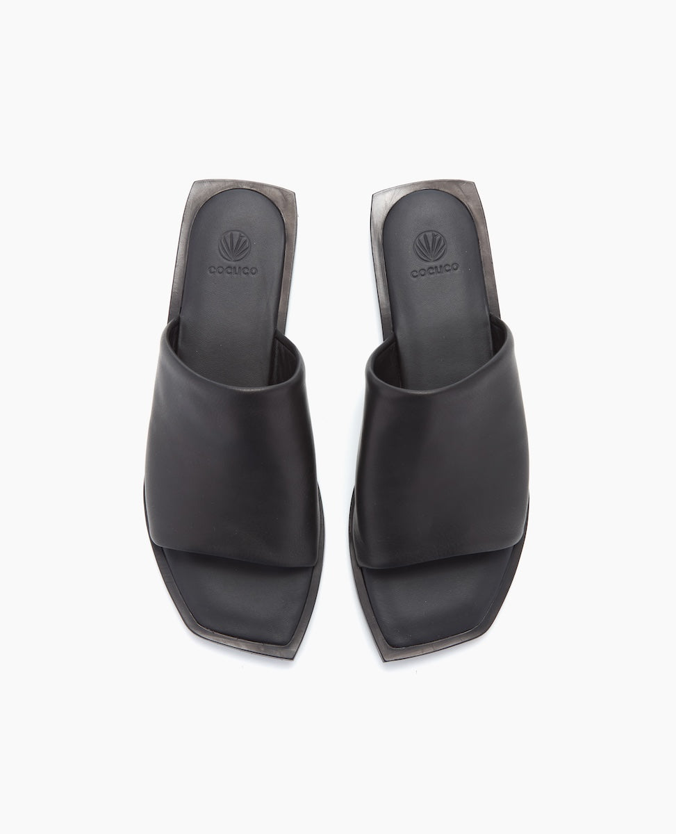 Sleek black sandals with a wide, smooth leather strap, featuring a square open toe design and a flat sole, embossed with a subtle logo on the insole.