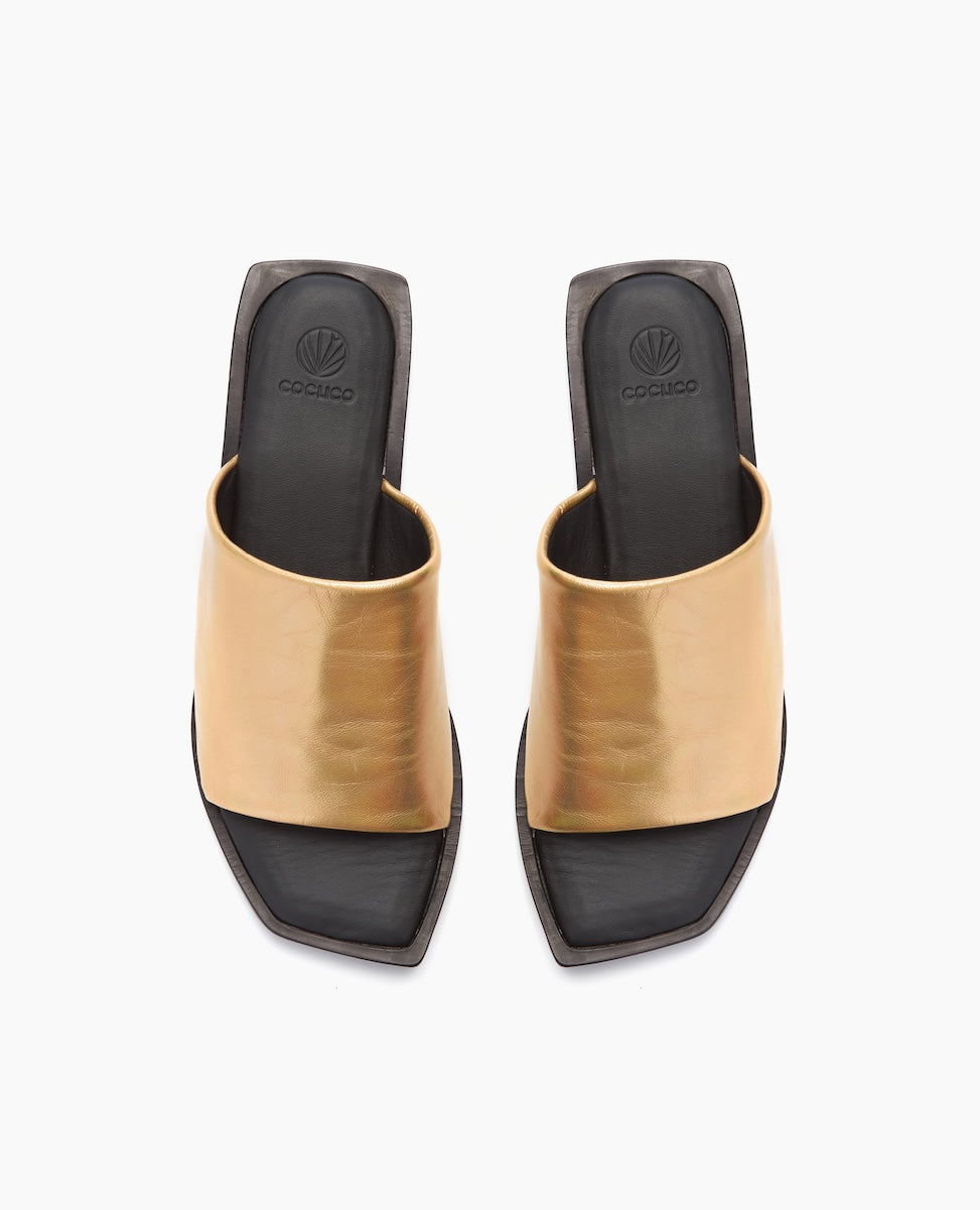 A pair of open-toed, square-heeled sandals featuring a wide, metallic gold strap across the top, with a sleek black footbed and a subtle logo embossed on the insole.