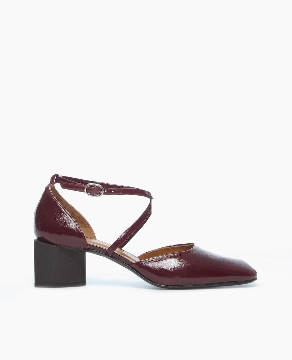 A burgundy patent leather shoe with a closed toe, block heel, and a cross-strap design featuring a small buckle.