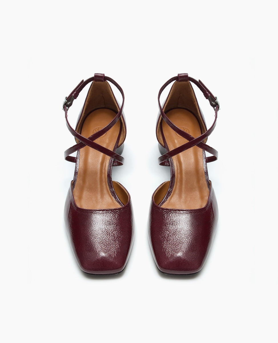 A pair of burgundy leather shoes with a glossy finish, featuring a square toe, crisscross ankle straps, and a medium heel.