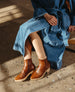 A person wearing a blue denim dress is seated, showcasing a pair of brown leather ankle boots with a smooth finish, tapered toes, and wooden platform heels. 10