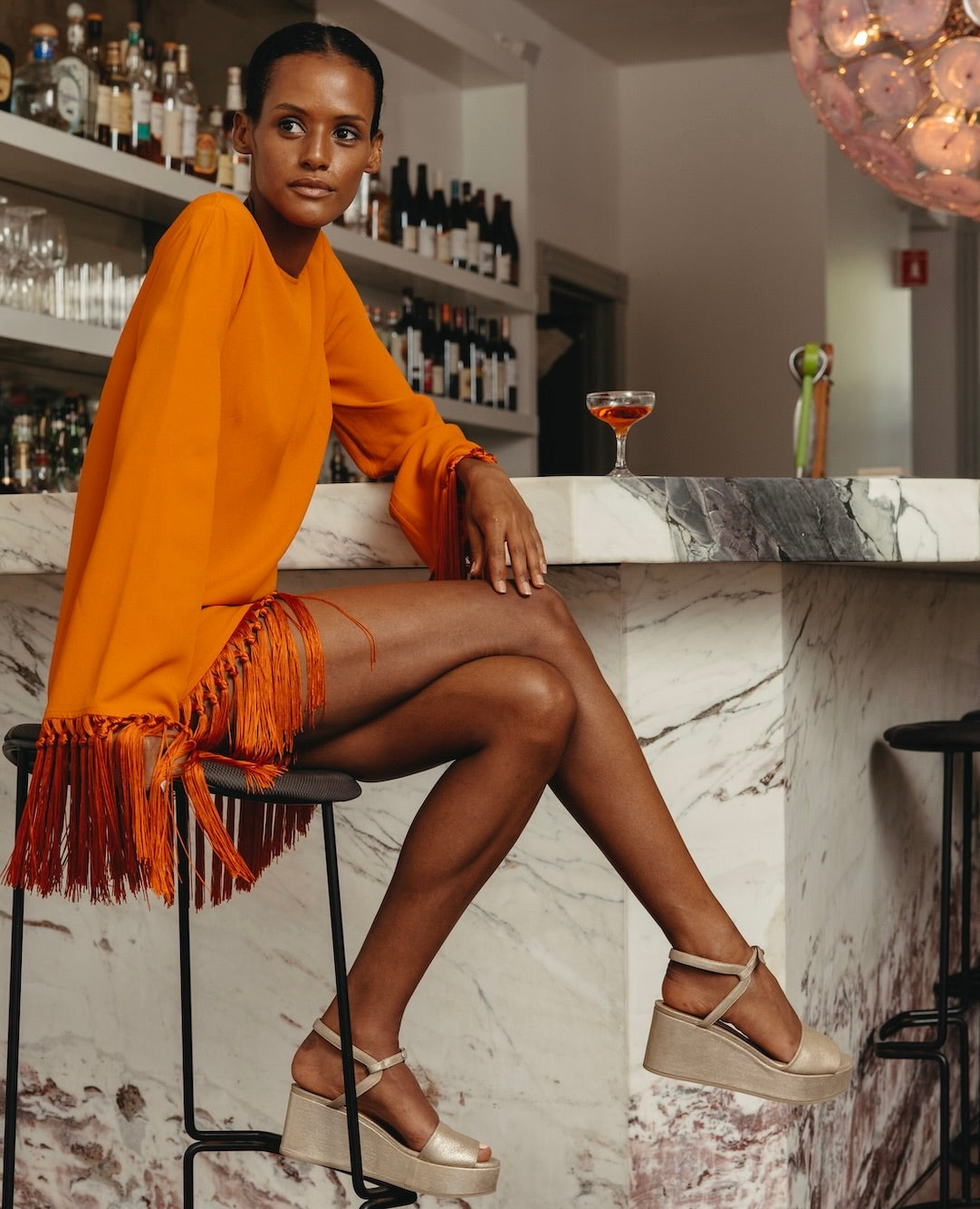 Woman seated at a bar wearing an orange fringe dress with her right leg crossed over her left and wearing the Lauren Wedge in Champagne Shimmer Suede.