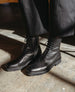 A person wearing black lace-up leather boots and black pants stands on a concrete floor. 10