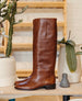 A tall, sleek, brown leather boot with a smooth texture, a rounded toe, a low block heel, and minimal stitching, standing upright on a wooden surface next to potted plants. 8