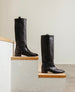 A pair of black knee-high leather boots is displayed on wooden steps against a minimalist background. 8