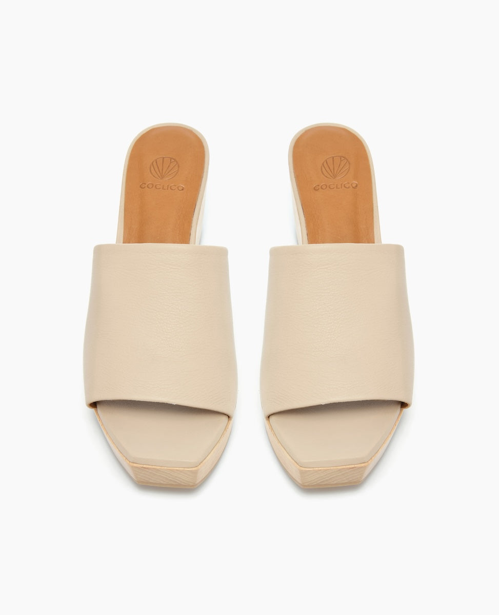 Beige slip-on sandals with a wide, smooth leather strap, open toe design, and a flat, square-shaped sole featuring a subtle logo imprint on the insole.