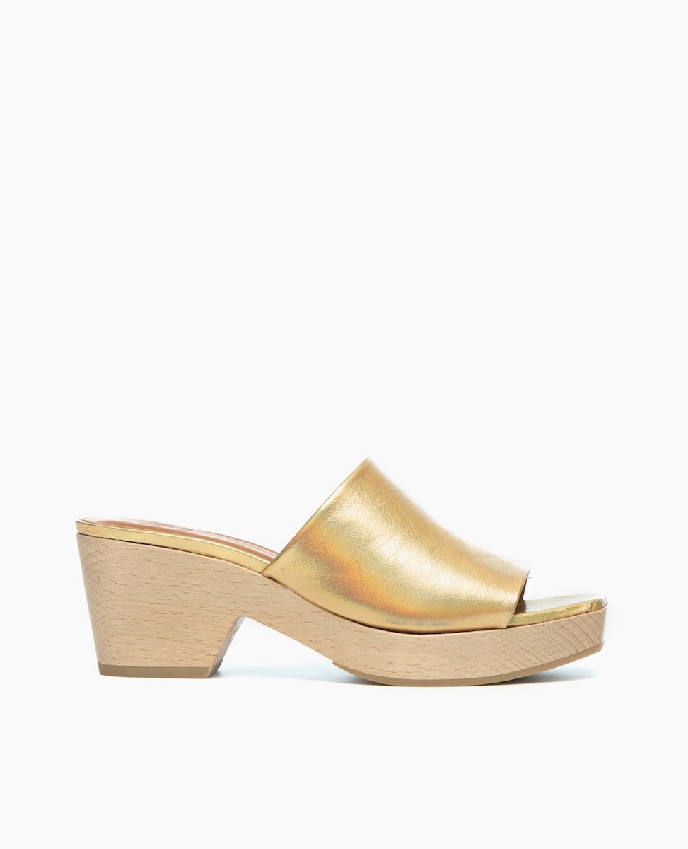 A gold, open-toe mule sandal with a smooth, metallic finish features a wide strap and a light wooden block heel.
