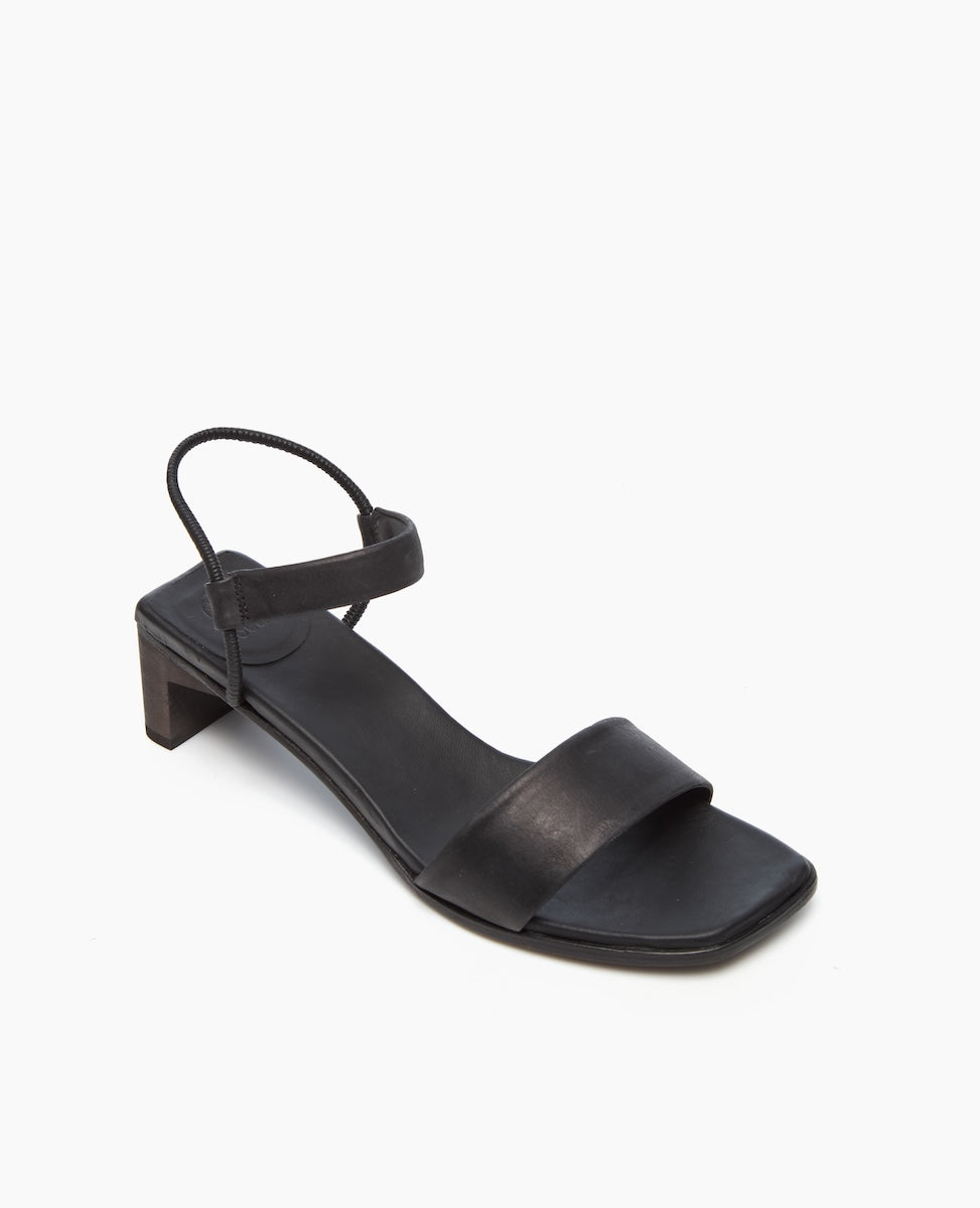 A sleek black sandal featuring a low block heel, a wide front strap, and a thin ankle strap with a minimalist design and smooth leather texture.