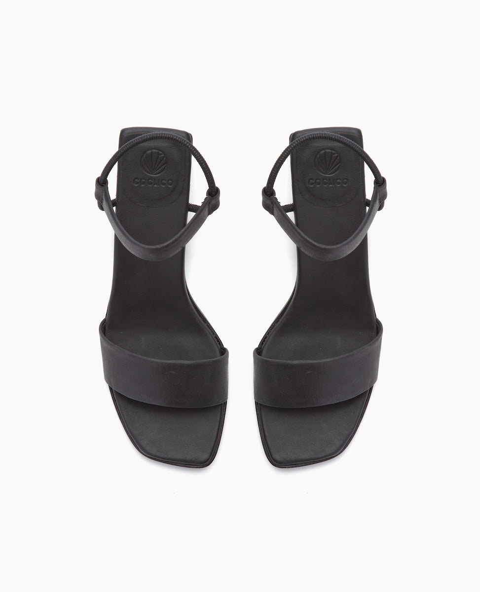 Black sandals with a minimalist design feature a smooth, matte finish, a single wide strap across the toes, an ankle strap with a buckle, and a flat sole.