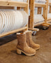 A pair of tan suede ankle boots with a side zipper, round toe, and chunky wooden heel, placed on a concrete floor near wooden shelves holding white ceramic plates. 7