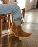 A pair of tan suede mid-calf boot with a wooden block heel, worn with light blue rolled-up jeans, featuring a rounded toe and a smooth, matte finish. 6