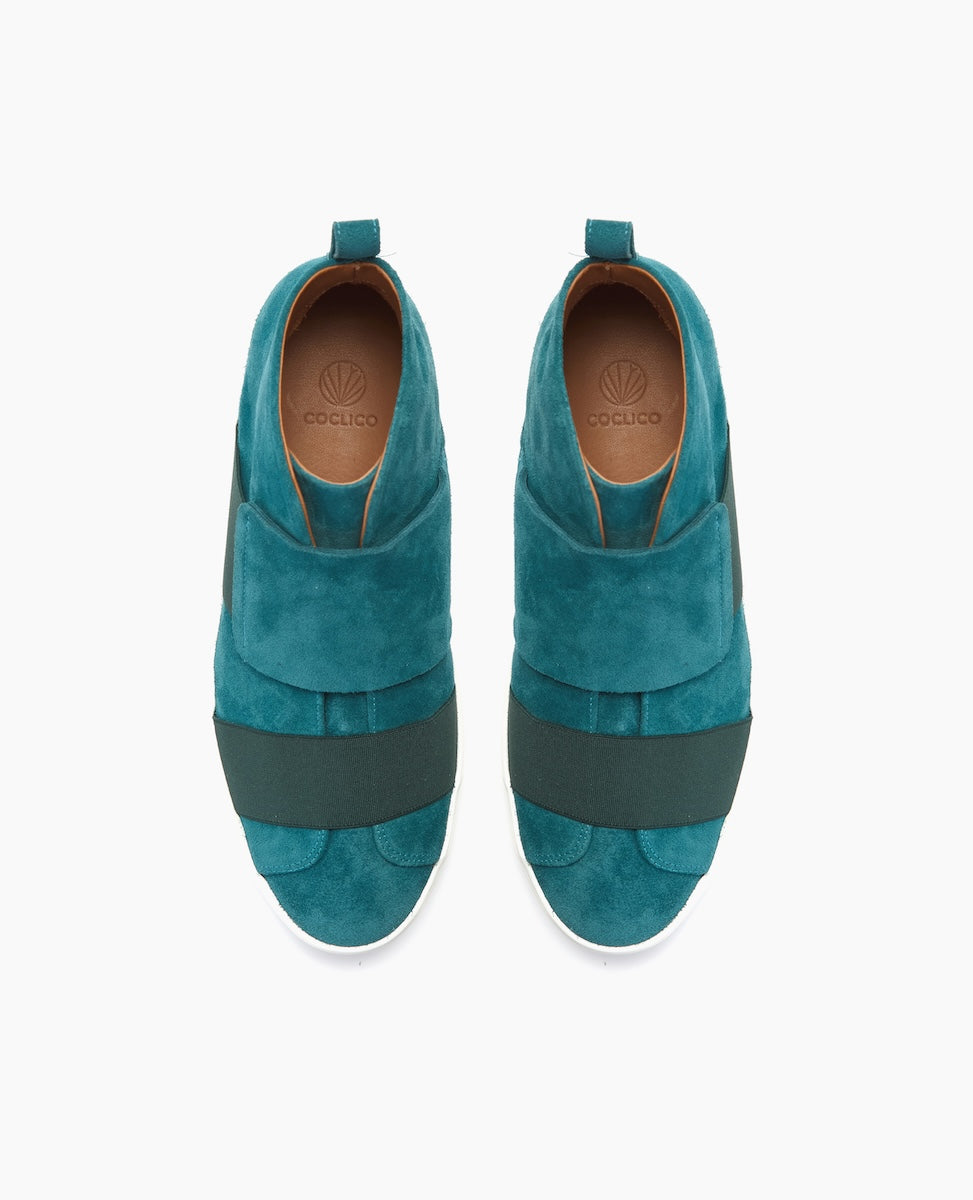 Teal suede slip-on shoes feature wide dark green elastic bands across the top, a pull tab at the heel, and a white rubber sole, with a smooth, rounded toe design.