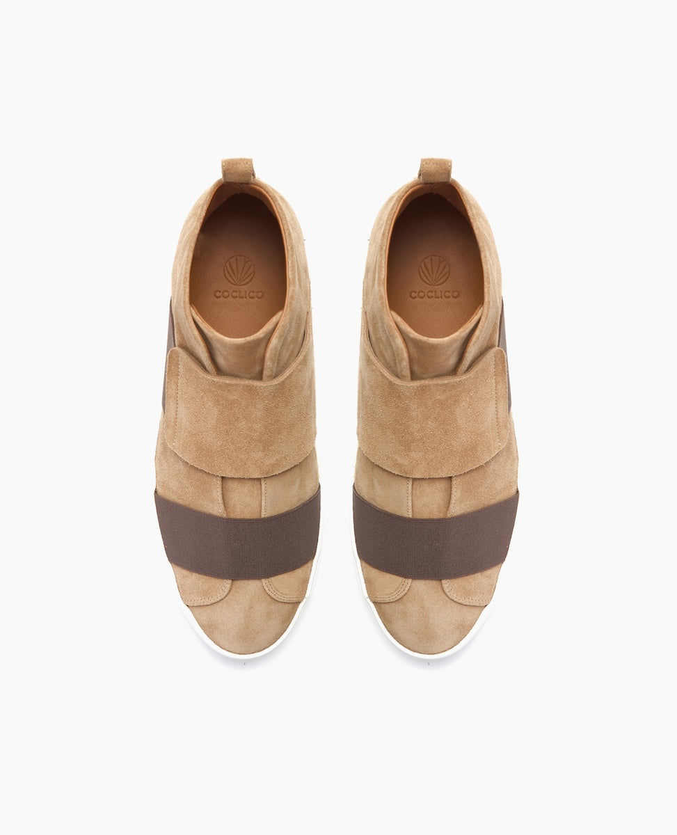 Tan suede slip-on shoes with wide brown elastic bands across the top, featuring a rounded toe, white rubber soles, and a pull tab at the heel.