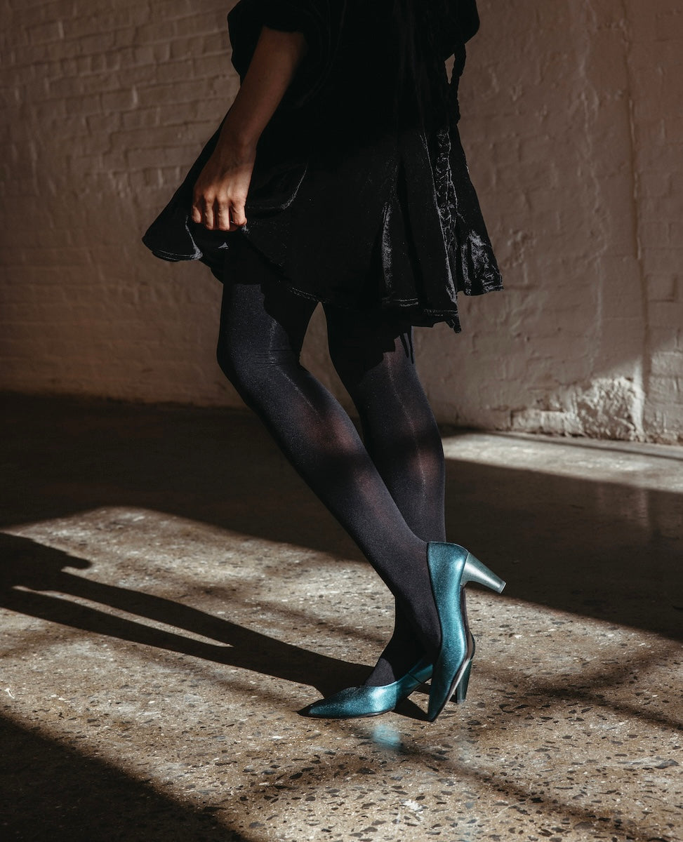 A person wearing teal high-heeled shoes with a pointed toe, black tights, and a black dress, standing on a polished concrete floor with a brick wall in the background.