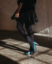 A person wearing teal high-heeled shoes with a pointed toe, black tights, and a black dress, standing on a polished concrete floor with a brick wall in the background. 5