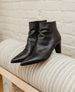 A pair of sleek, black leather ankle boots with pointed toes, a smooth texture, and a moderate heel, displayed on a light-colored, ribbed surface against a brick wall background. 10