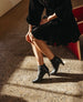 A person wearing a black, long-sleeved dress with a ruffled hem and black ankle boots sits on a red bench, with sunlight casting shadows on the speckled floor. 12