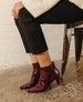 A pair of sleek, burgundy ankle boots with a glossy, textured finish, pointed toes, and oval hand painted heel, worn with black pants and a white sweater. 11