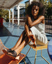 Woman seated outside on a yellow chair with her legs and feet rested  on an orange chair and wearing the Sokolo Heel in Black Leather. 6